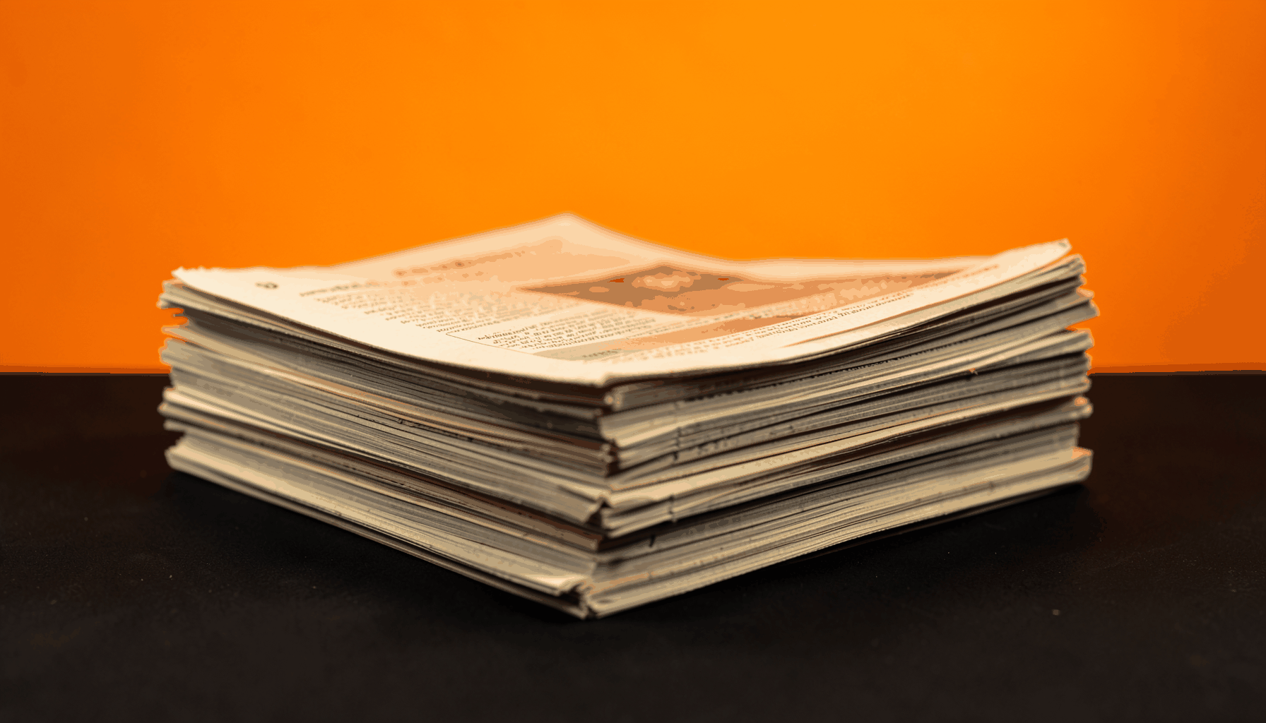 A stack of neatly folded newspapers on a plain charcoal surface, lit softly in warm gold tones, calm editorial mood