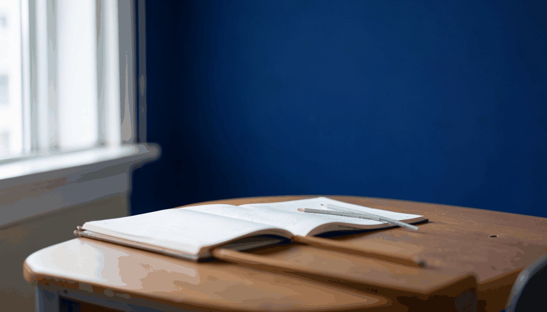 A quiet classroom desk with an open notebook and pencil, soft natural light from a window, calm and studious atmosphere